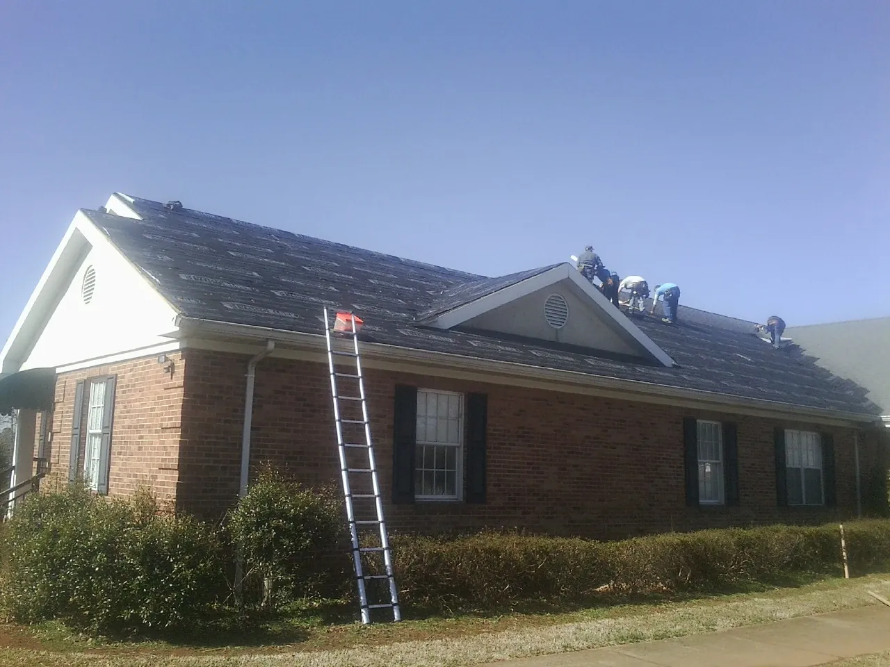 Crew tearing shingles off a brick ranch roof with a ladder set — Emergency Roof Repair work by our crew in Pecatonica