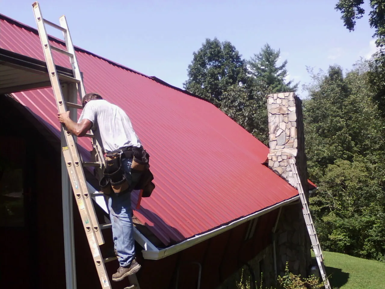 Roofer on a ladder beside a red metal roof and stone chimney — Soffit & Fascia Repair by Roofing Brothers in Pecatonica
